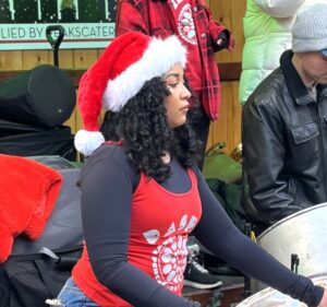 Girl with curly hair and Christmas hat plays steel pans - Aman wears Christmas hat and plays guitar pans close up - RASPO Calypso & Carols 2025 image by Mary Genis