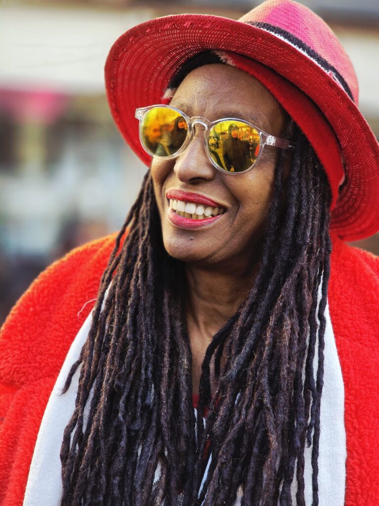 A black woman with red lipstick and dreadlocks smiles wearing a red had and coat with reflective sunglasses - RASPO Calypso & Carols 2025 image by Julia Brazil