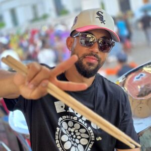 Image of drummer and drum tutor Jamah holding drum sticks wearing a black and silver RASPO steel band t-shirt