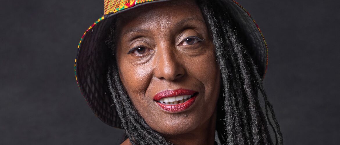 Image of Mary Genis a black woman with locks wearing an kente print hat and smiling into the camera